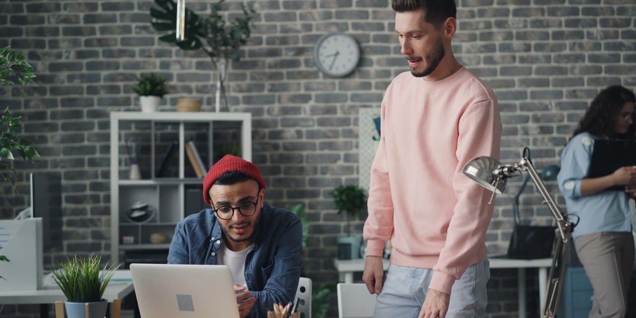 Developer in a red beanie works on a laptop while a colleague stands at the desk checking his work, in a brick-walled office
